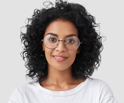 Close up portrait of curly female adult has charming smile, curly dark hair, wears big glasses, satisfied as finished domestic work earlier, being successful designer or architect, has talent