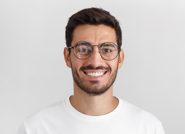 Daylight portrait of young handsome caucasian man isolated on grey background, dressed in white t-shirt and round eyeglasses, looking at camera and smiling positively